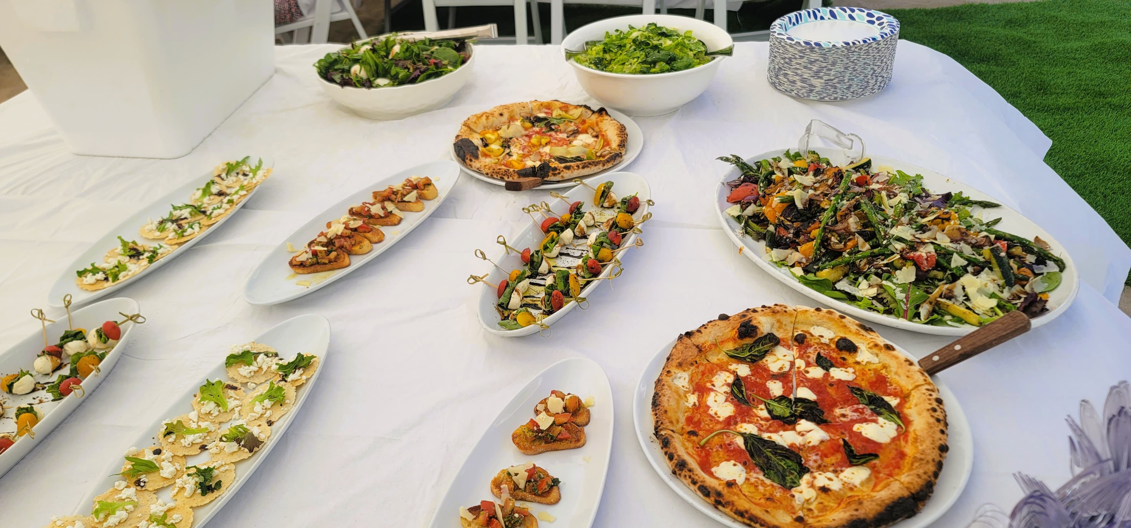 Buffet spread with pizzas, caprese skewers, bruschetta, and salads on white platters