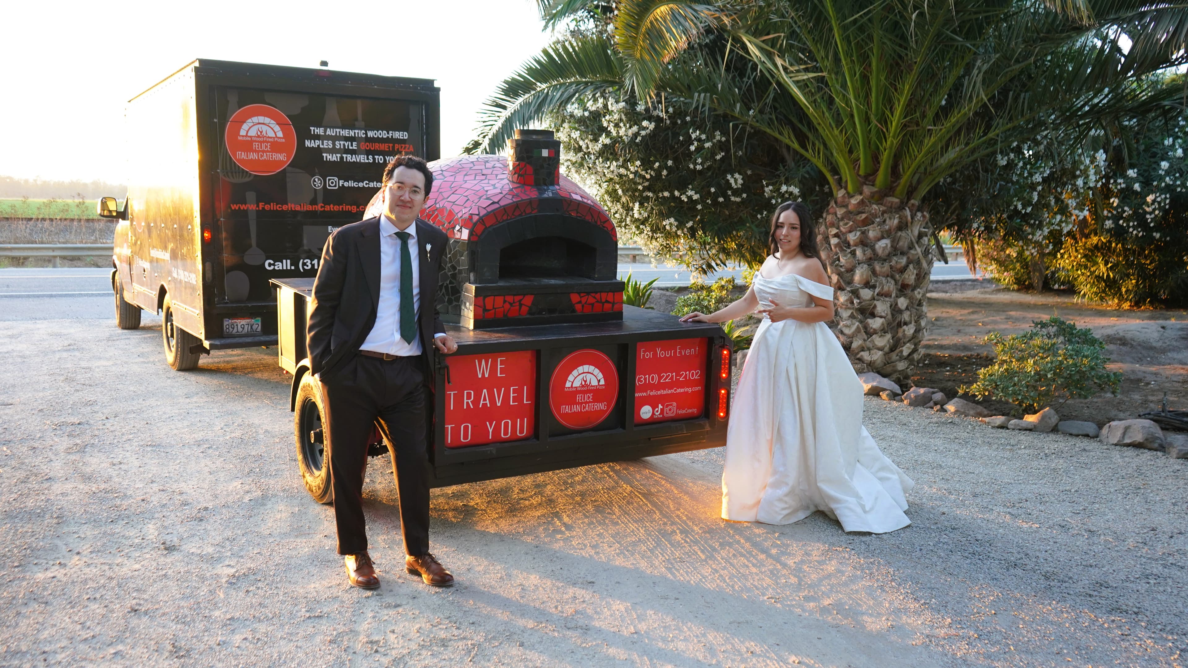 Newlyweds standing beside the Felice wood-fired pizza trailer at their wedding